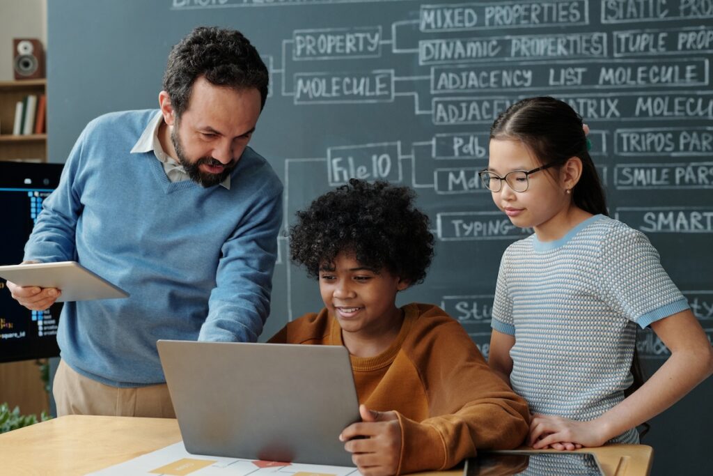 Teacher offering guidance to two focused students working on laptop during class. Blackboard with scientific terms visible in background showcasing an educational environment